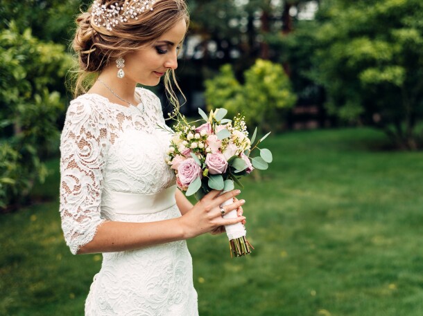 the bride in a white long dress with a wedding bouquet is standing alone and looks at a bouquet of flowers