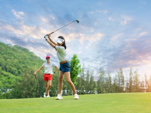 Golf ball on tee ready to be struck by woman golfer as partner watches with sunset
